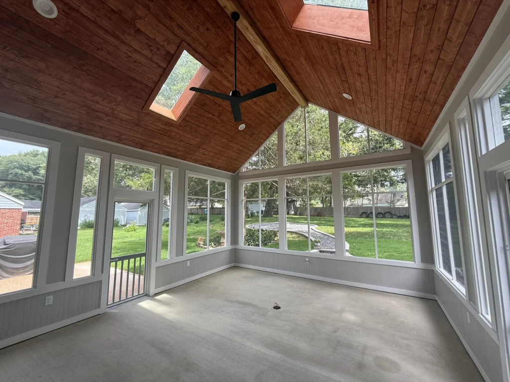 Mostly completed interior of a screened-in porch addition with a concrete floor, wood-stained ceiling, two skylights, black ceiling fan, and two doors.